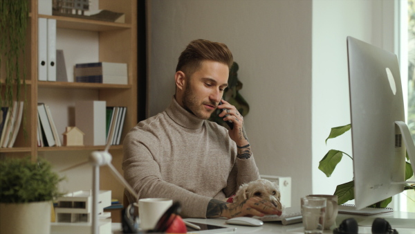 A young businessman with dog sitting at the desk indoors in office, using smartphone and computer.