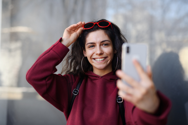 A portrait of young woman with smartphone standing outdoors on street in city, taking selfie.