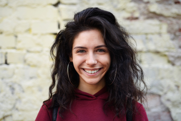 Portrait of happy young woman standing outdoors against stone background, looking at camera.