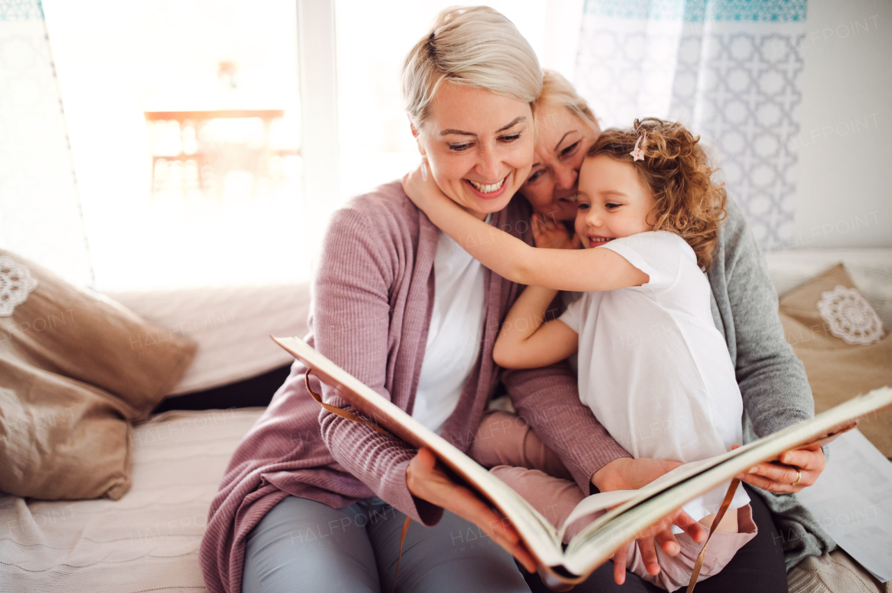 A small girl with mother and grandmother sitting on a sofa at home, looking at photographs.