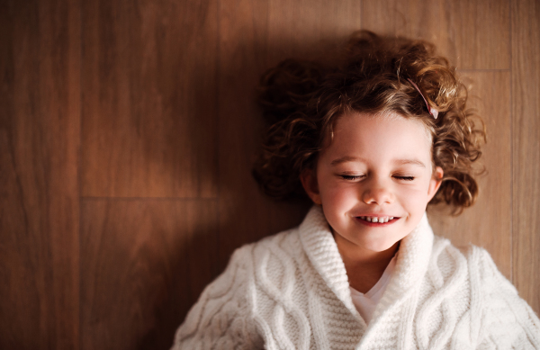 A close-up portrait of happy small girl with white knitted sweater lying on the floor, a top view.