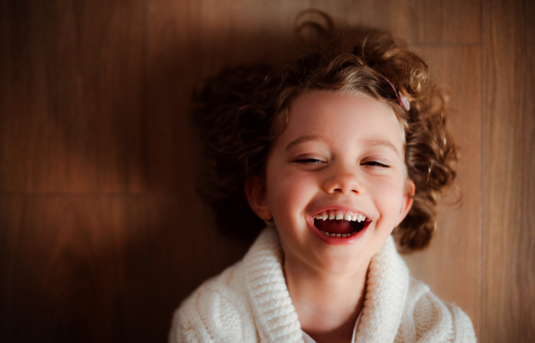 A close-up portrait of happy small girl with white knitted sweater lying on the floor, a top view.