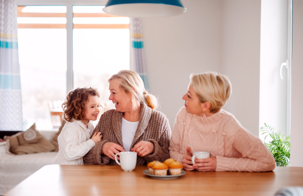 A portrait of happy small girl with mother and grandmother sitting at the table at home, eating muffins.