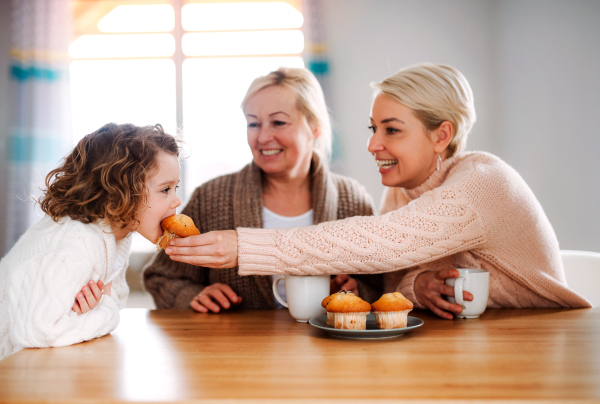 A portrait of happy small girl with mother and grandmother sitting at the table at home, eating muffins.