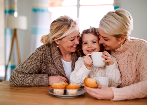 A portrait of happy small girl with mother and grandmother sitting at the table at home, drinking.