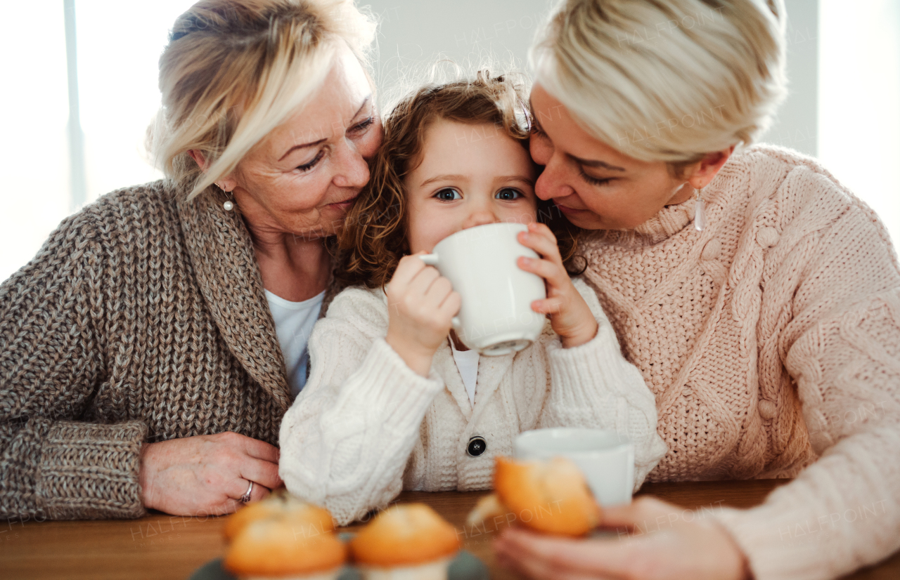 A portrait of happy small girl with mother and grandmother sitting at the table at home, drinking.