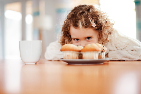 A portrait of happy small girl with muffins sitting at the table at home.