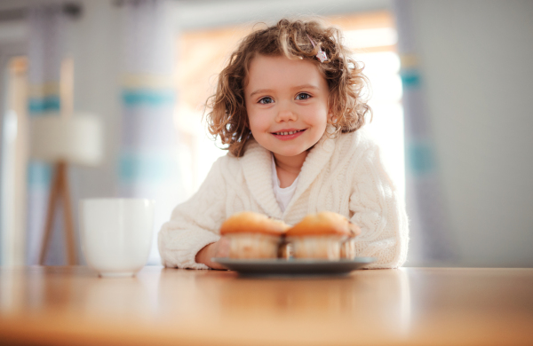 A portrait of happy small girl with muffins sitting at the table at home.