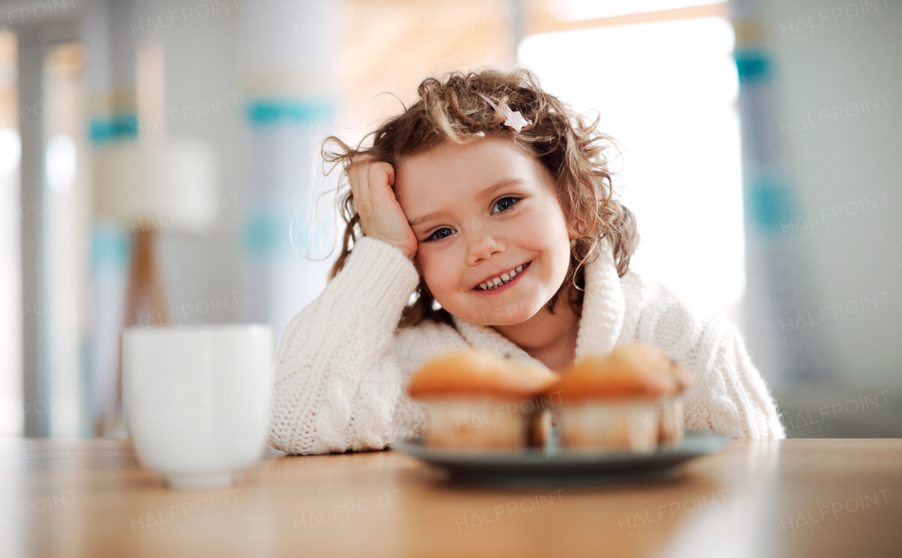 A portrait of happy small girl sitting at the table at home, eating muffins.