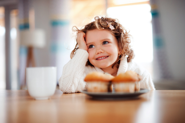 A portrait of happy small girl with muffins sitting at the table at home.