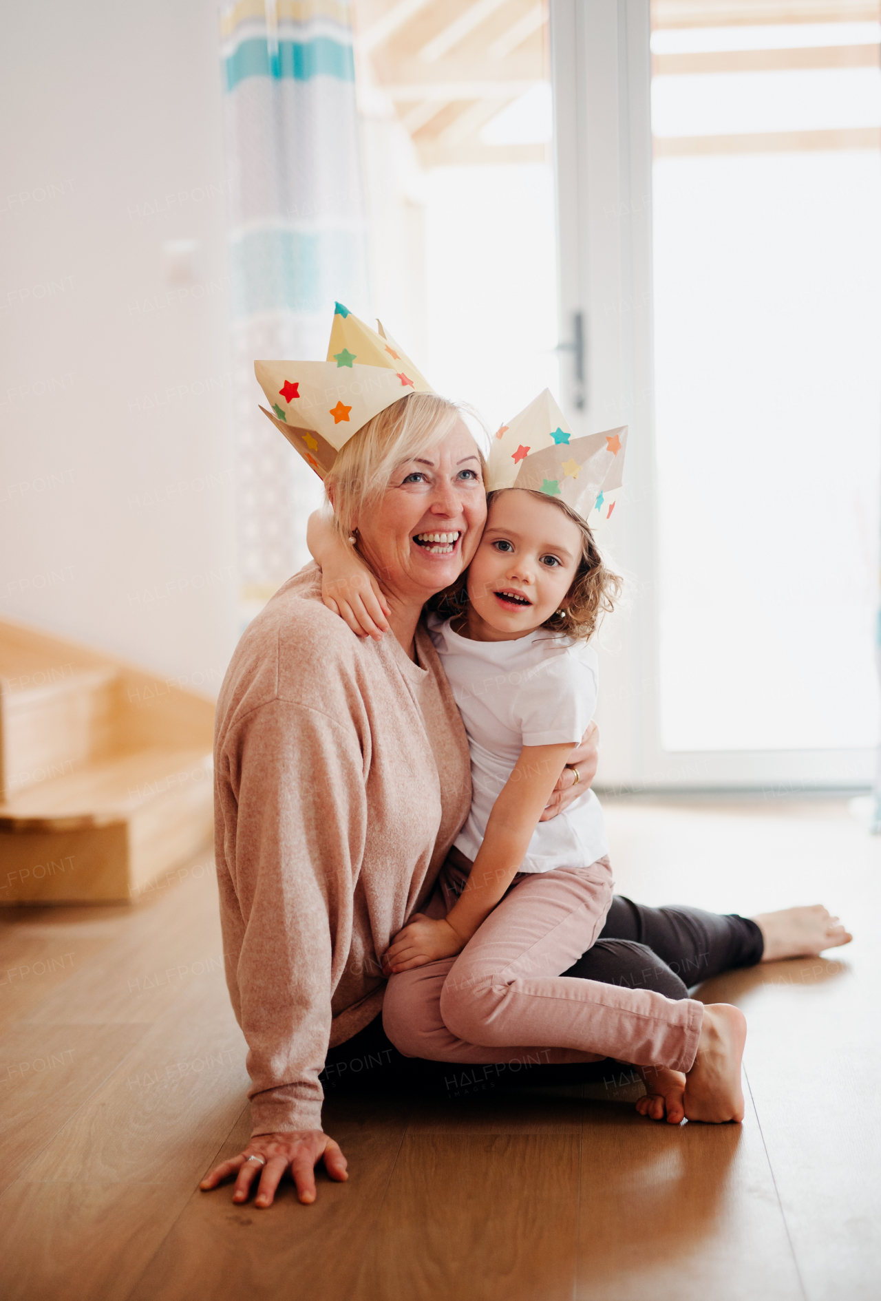 A portrait of small girl and grandmother with paper crown hugging at home.