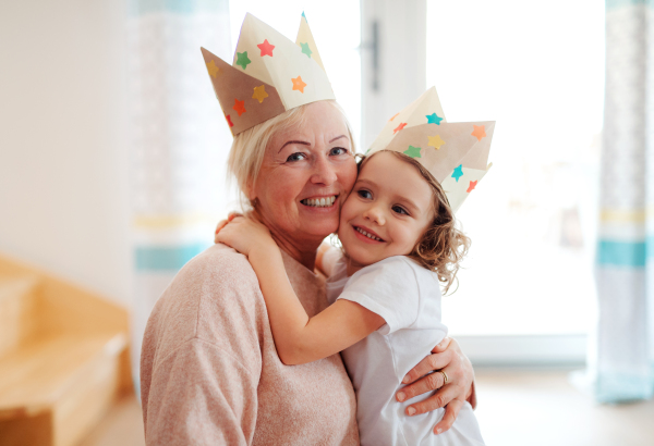 A portrait of small girl and grandmother with paper crown hugging at home.