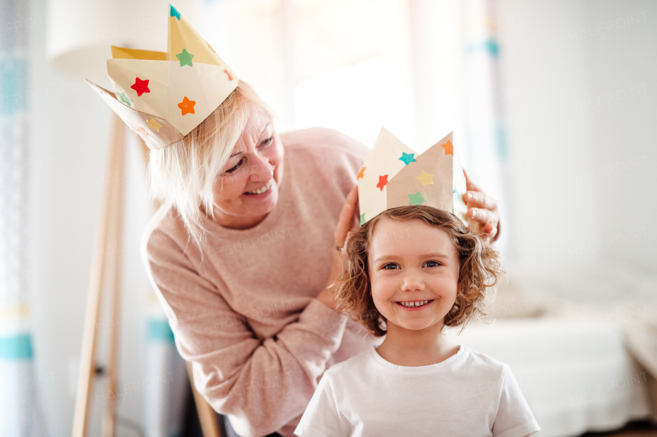 A portrait of small girl and grandmother with paper crown at home, having fun.