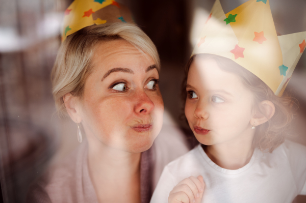 A portrait of small girl and mother with paper crown having fun at home. Shot through glass.