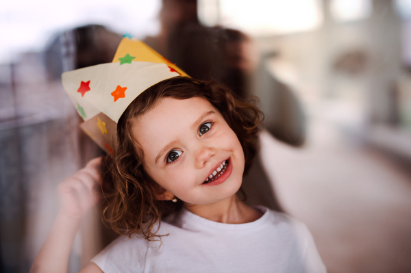 A portrait of small girl with a paper crown at home, looking at camera. Shot through glass.