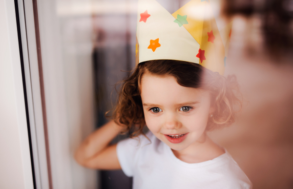 A portrait of small girl with paper crown on head at home, looking out of a window. Shot through glass.