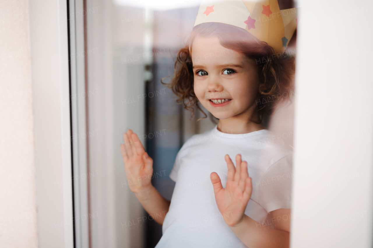 A portrait of small girl with paper crown on head at home, looking out of a window. Shot through glass.