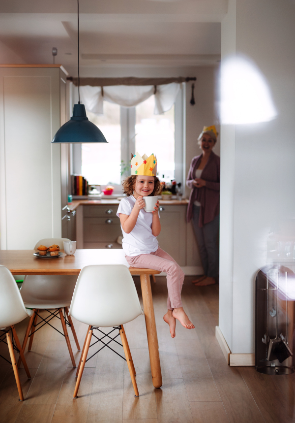 A side view portrait of small girl and mother with paper crown at home, drinking.