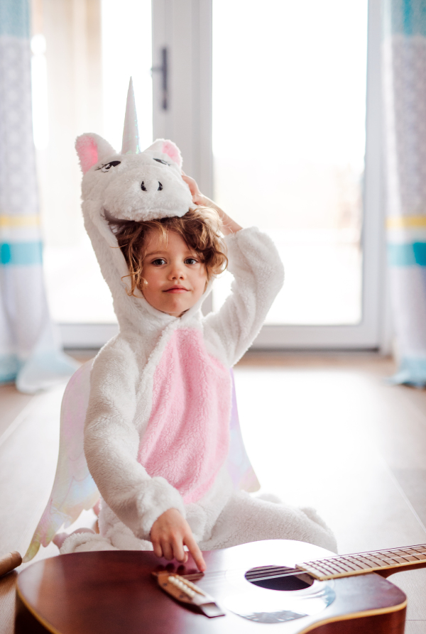 A portrait of happy small girl in unicorn mask with guitar at home.