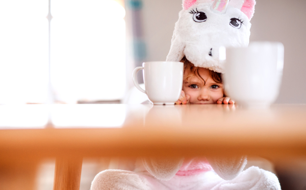 A portrait of happy small girl in unicorn mask sitting at the table at home.