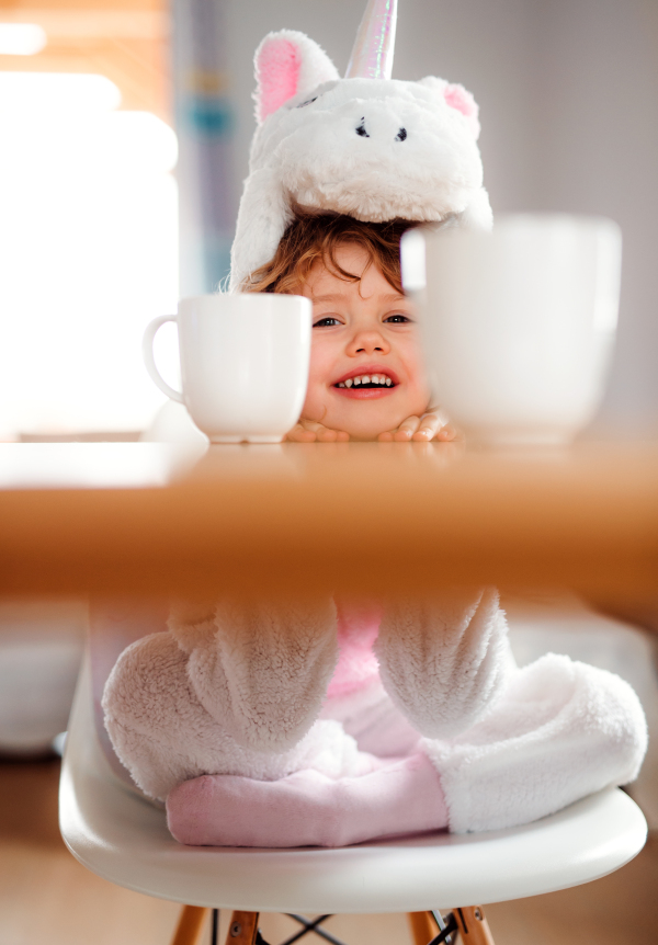 A portrait of happy small girl in unicorn mask sitting at the table at home.