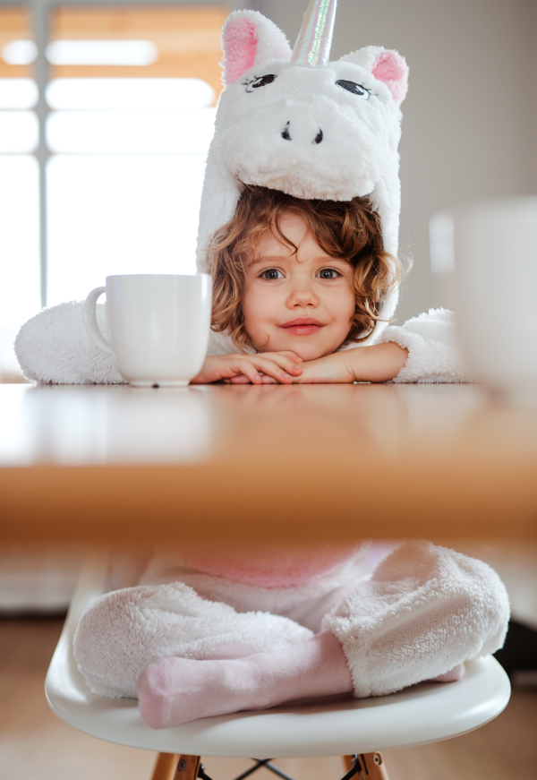 A portrait of happy small girl in unicorn mask sitting at the table at home.