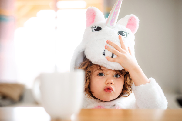 A portrait of happy small girl in unicorn mask sitting at the table at home.