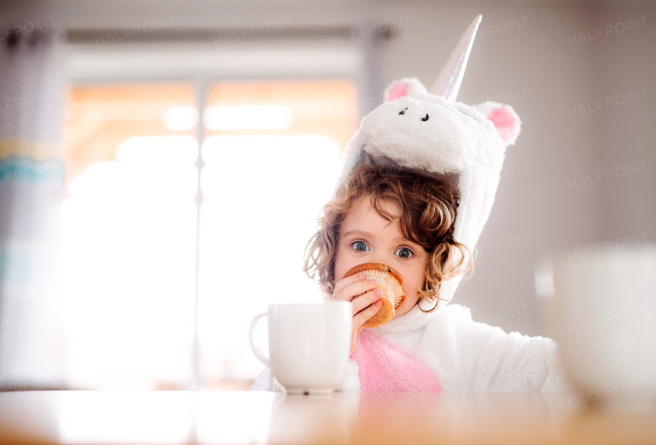 A portrait of happy small girl in unicorn mask sitting at the table at home, eating muffin.