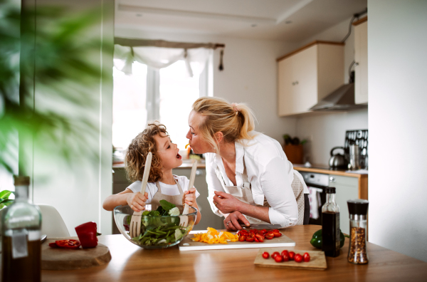 A portrait of small girl with grandmother in a kitchen at home, preparing vegetable salad.