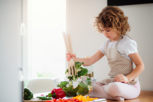 A portrait of small girl in a kitchen at home, preparing vegetable salad.