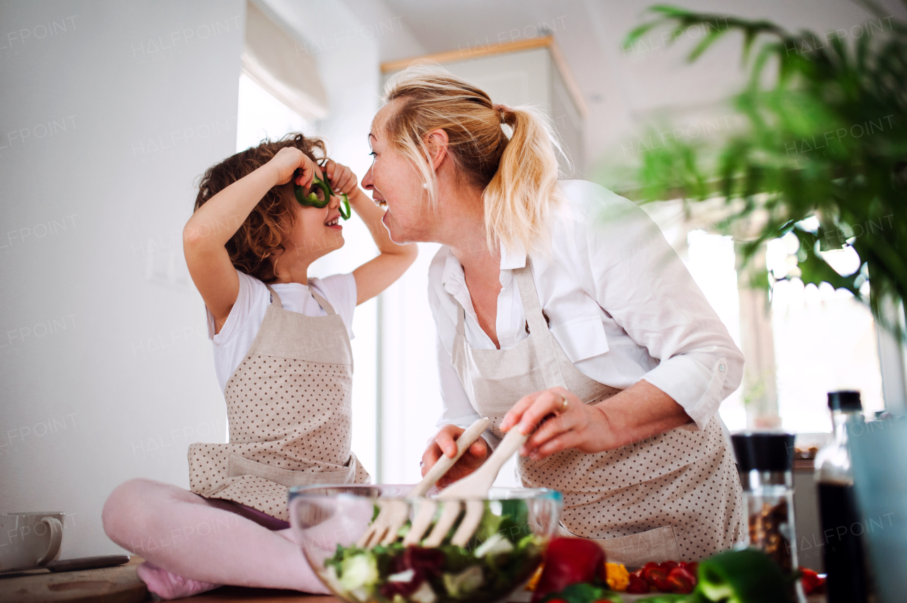 A portrait of happy small girl with grandmother preparing vegetable salad at home, having fun.