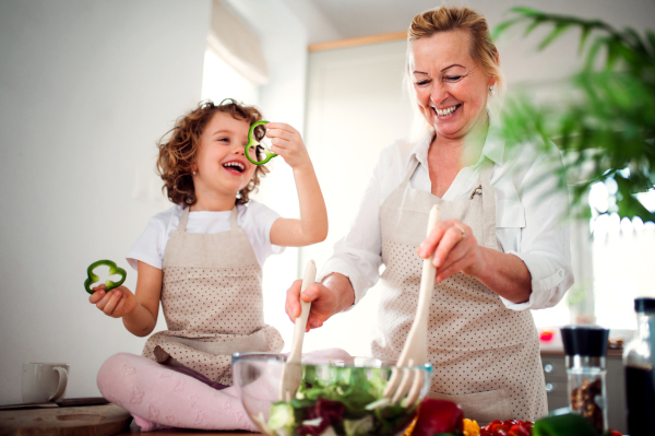 A portrait of small girl with grandmother in a kitchen at home, preparing vegetable salad.