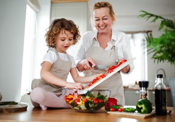 A portrait of small girl with grandmother in a kitchen at home, preparing vegetable salad.