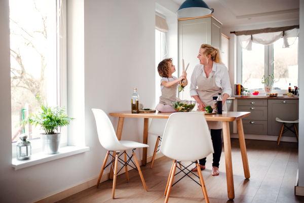 A portrait of small girl with grandmother in a kitchen at home, preparing vegetable salad.