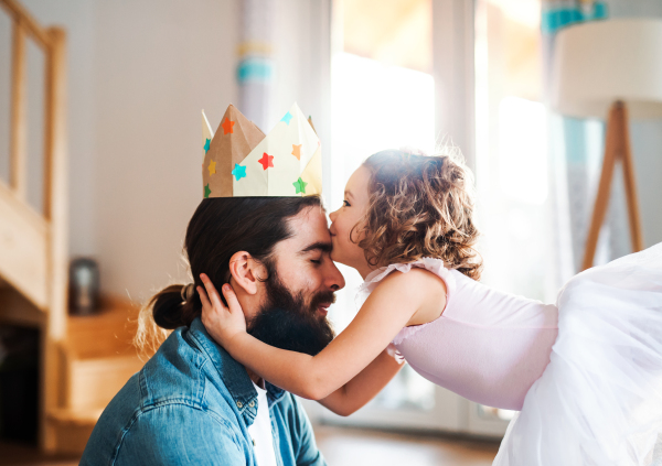 A side view of small girl with a princess crown and young father at home, kissing when playing.