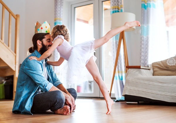 A side view of small girl with a princess crown and young father at home, playing.