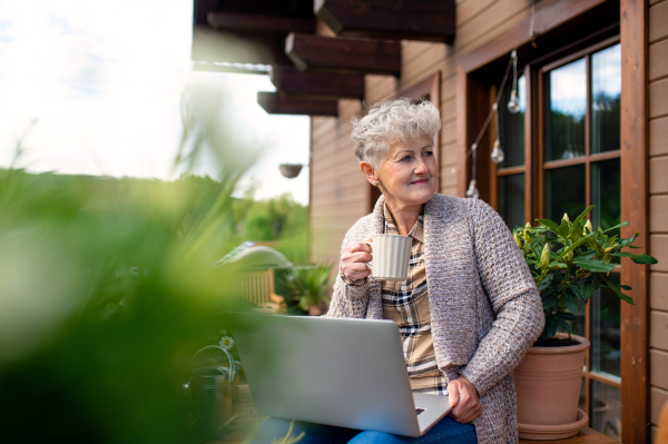 Portrait of senior woman with laptop and coffee sitting on terrace in summer, resting.