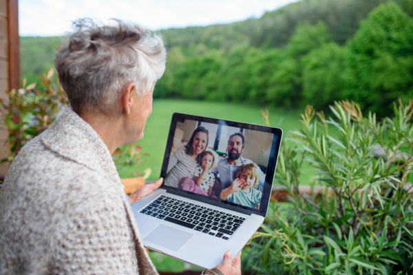 Senior woman with laptop sitting on terrace in summer, having video call with family.