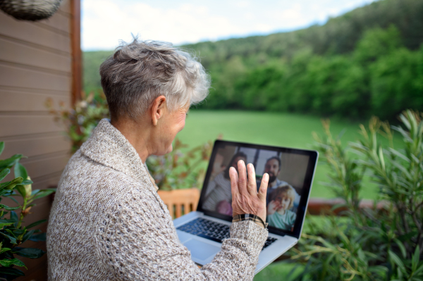 Senior woman with laptop sitting on terrace in summer, having video call with family.