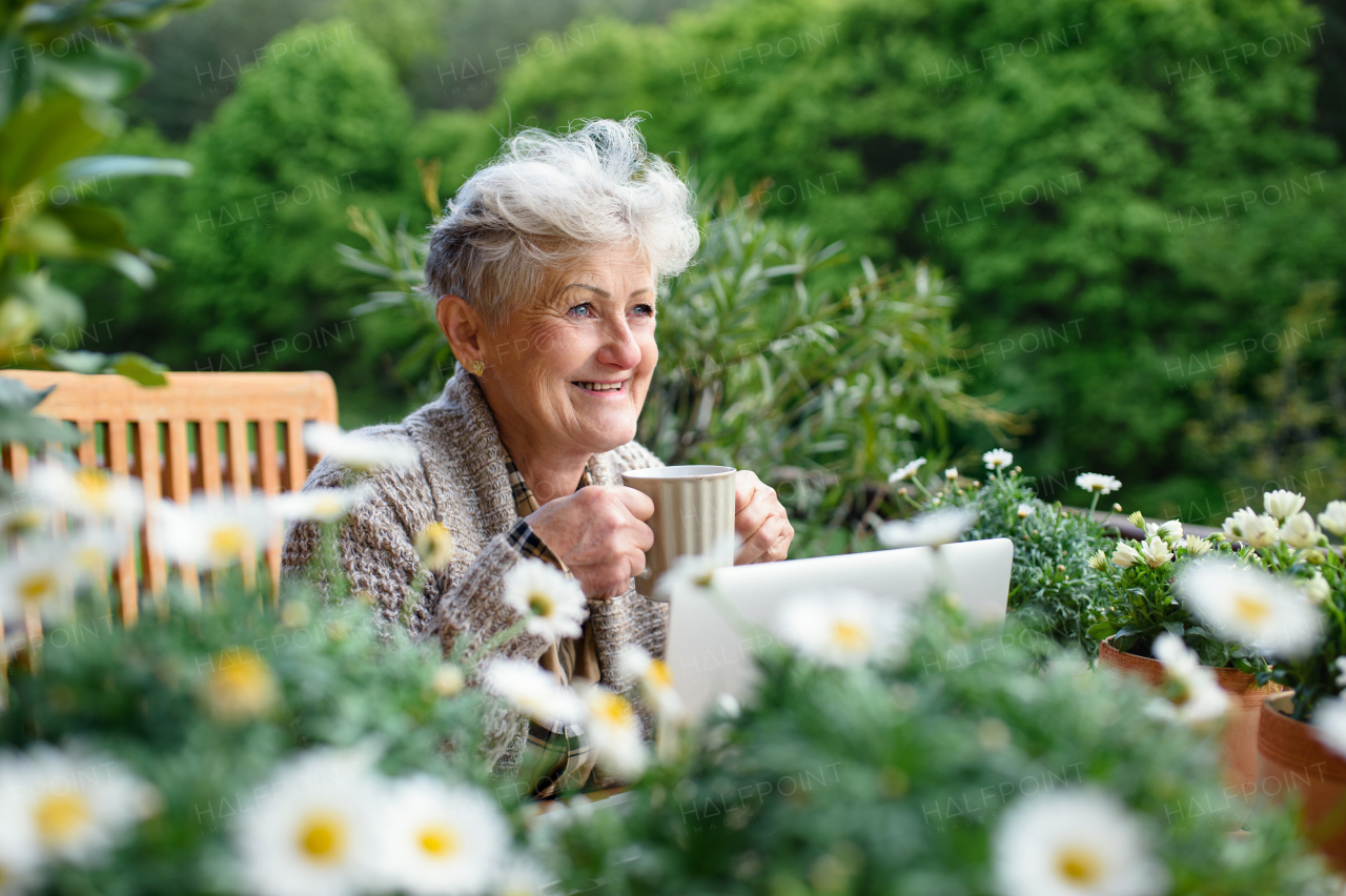 Portrait of senior woman with laptop and coffee sitting on terrace in summer, resting.