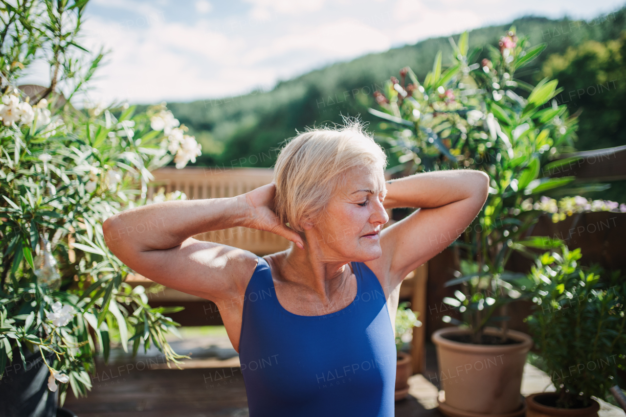 A senior woman standing outdoors on a terrace in summer, doing stretching. Copy space.