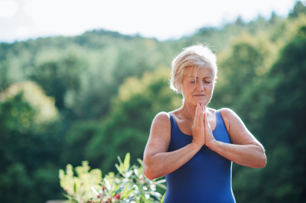 A senior woman standing outdoors on a terrace in summer, doing yoga exercise. Copy space.