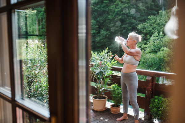 A senior woman with sports bra outdoors on a terrace in summer, drinking water. Shot through glass.