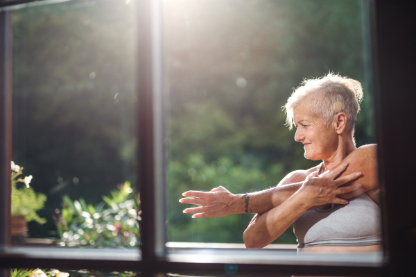 A front view of senior woman with sports bra standing outdoors on a terrace in summer, stretching.