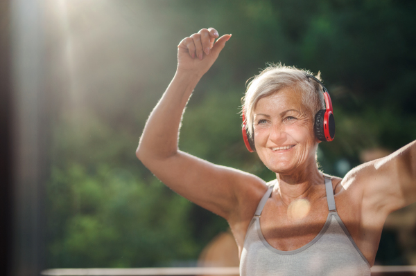 A senior woman with headphones standing outdoors on a terrace in summer, listening to music.