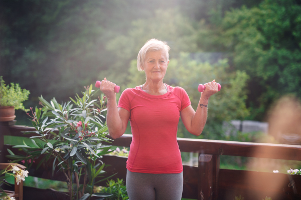 A senior woman standing outdoors on a terrace in summer, doing exercise with dumbbells. Copy space.