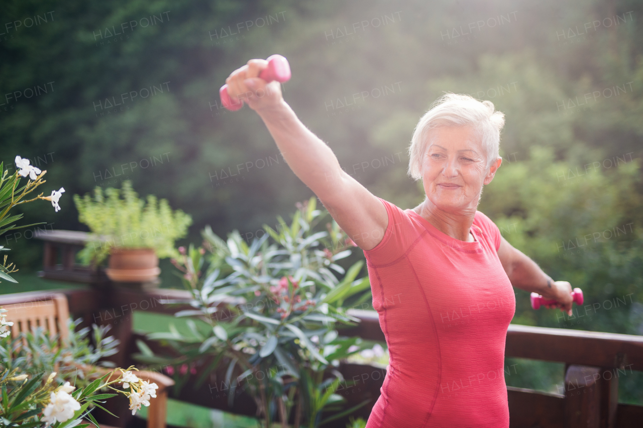 A senior woman standing outdoors on a terrace in summer, doing exercise with dumbbells. Copy space.