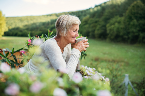 A senior woman with coffee standing outdoors on a terrace in summer.