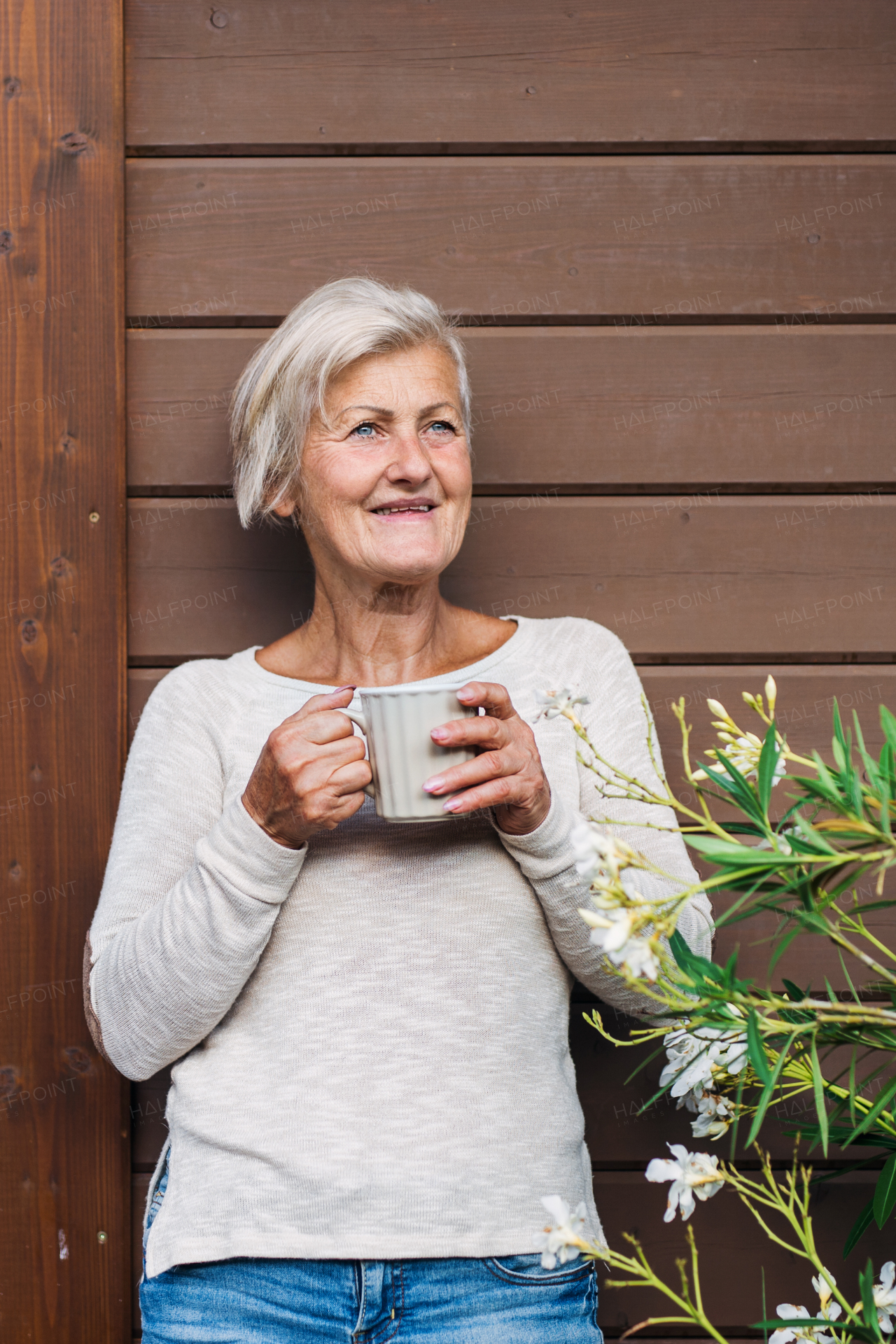 A happy senior woman with coffee standing outdoors on a terrace in summer.