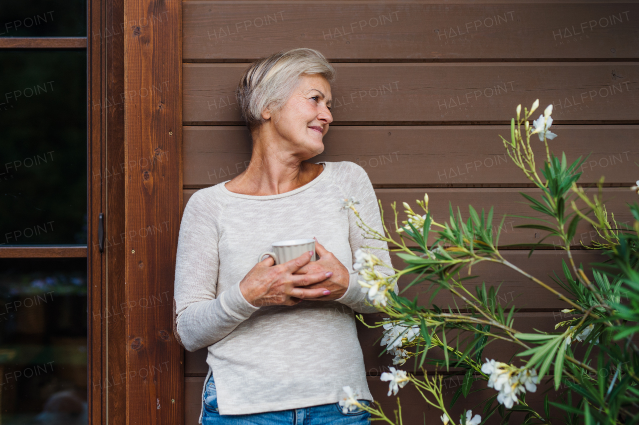 A happy senior woman with coffee standing outdoors on a terrace in summer.